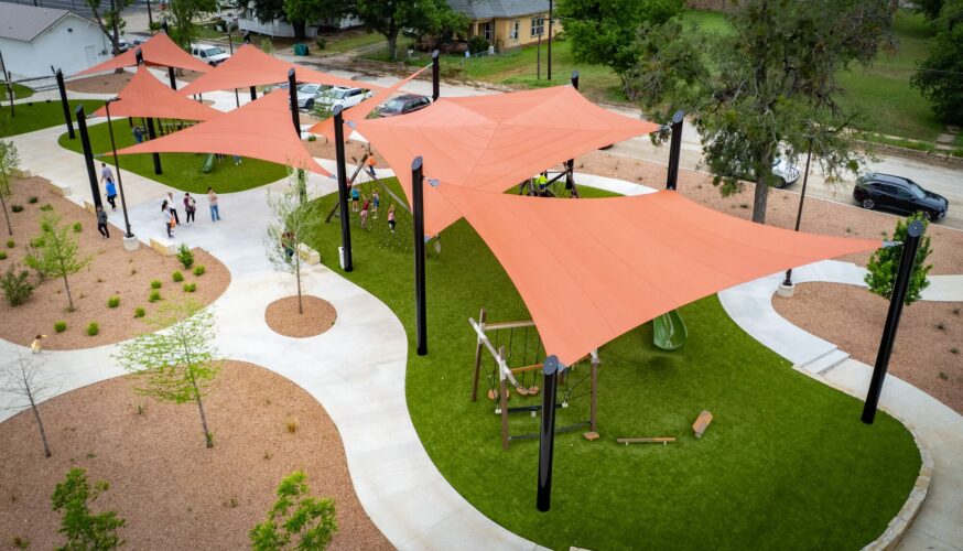 Children playing at a playground under red shade sails at District 1876, Coleman, Texas.