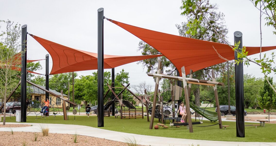 Children playing at a playground under red shade sails at District 1876, Coleman, Texas.