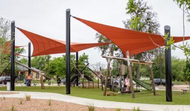 Children playing at a playground under red shade sails at District 1876, Coleman, Texas.