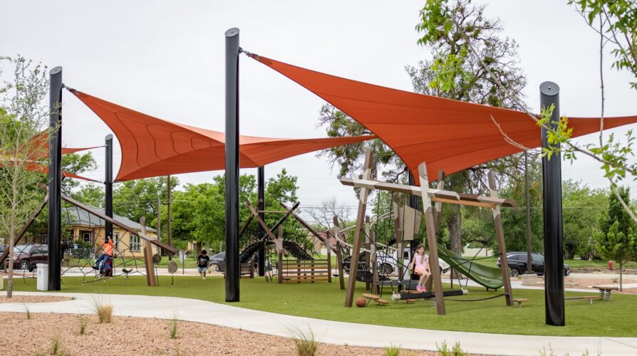 Children playing at a playground under red shade sails at District 1876, Coleman, Texas.