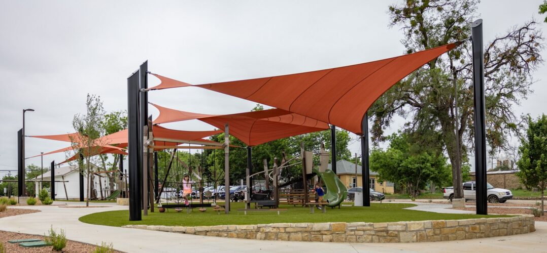 Children playing at a playground under red shade sails at District 1876, Coleman, Texas.