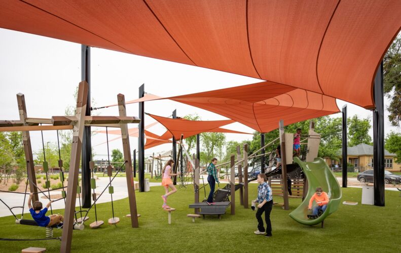 Children playing at a playground under red shade sails at District 1876, Coleman, Texas.