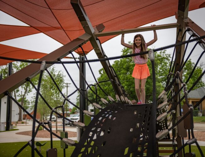 Children playing at a playground under red shade sails at District 1876, Coleman, Texas.