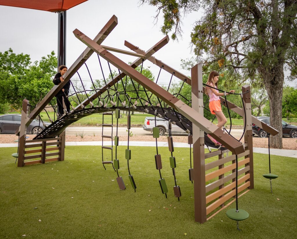 Children playing at a playground under red shade sails at District 1876, Coleman, Texas.